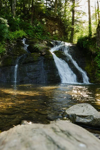 Temiz bir havuza dökülen bir orman şelalesinin sakin dikey görüntüsü. Düşük açılı perspektif ve güneşli kayalar doğa için barışçıl bir atmosferi çağrıştırıyor.