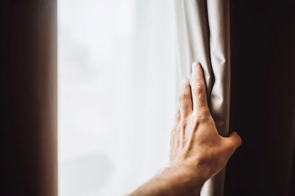 A hand pushes the blackout curtains away from the window - bright morning light of a new day