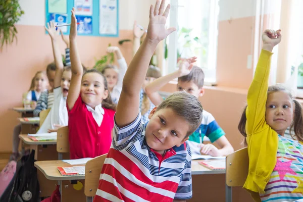 Niños en escritorios con las manos levantadas —  Fotos de Stock