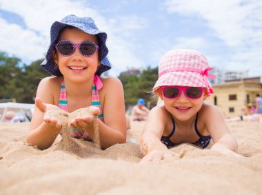 Küçük kızlar kumsalda oynuyorlar ve güneşleniyorlar.