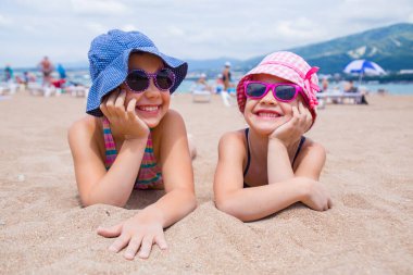 Küçük kızlar kumsalda oynuyorlar ve güneşleniyorlar.