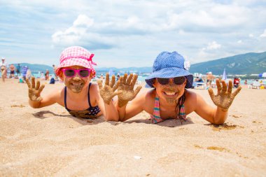 Küçük kızlar kumsalda oynuyorlar ve güneşleniyorlar.