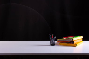Kalem saksısı istiflenmiş kitapların yanında, okul malzemeleri beyaz masada, arka planda karatahta deseni var. Yan görüş, kopyalama alanı. Öğrenme, eğitim kavramı 
