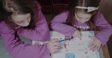 Pembe kazak giyen iki kız kardeş ödevlerini masa renkleri ve kalemleriyle yapıyorlar.