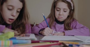 Pembe kazak giyen iki kız kardeş ödevlerini deftere, masa renklerine ve kalemlere yazıyorlar.