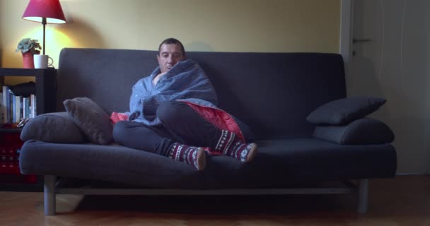 Ένας Άντρας Πυρετό Που Κάθεται Στον Καναπέ Τυλιγμένος Μπλε Κουβέρτα — Αρχείο Βίντεο