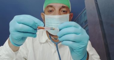 Coronavirus aşısı laboratuvar testi sırasında şırınga kullanan beyaz önlük, cerrahi maske, mavi eldiven ve tıbbi başlık giyen bir doktor görülüyor.