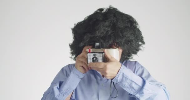 一个有着卷曲毛茸茸的镜头的男人的前视图 他带着一台被灰色背景隔离的复古即时相机 — 图库视频影像