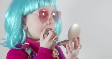 Güneş gözlüğü takan ve mavi peruklu sevimli küçük kız dudaklarını rujla boyuyor, küçük aynanın önünde makyaj yapmayı öğreniyor. Yetişkinlerin kozmetiklerle deney yapmalarını taklit eder