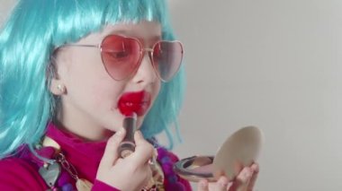 Güneş gözlüğü takan ve mavi peruklu sevimli küçük kız dudaklarını rujla boyuyor, küçük aynanın önünde makyaj yapmayı öğreniyor. Yetişkinlerin kozmetiklerle deney yapmalarını taklit eder