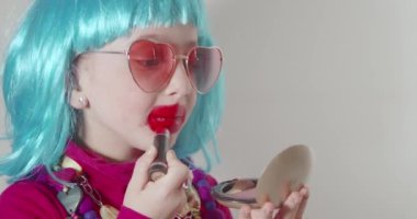 Güneş gözlüğü takan ve mavi peruklu sevimli küçük kız dudaklarını rujla boyuyor, küçük aynanın önünde makyaj yapmayı öğreniyor. Yetişkinlerin kozmetiklerle deney yapmalarını taklit eder