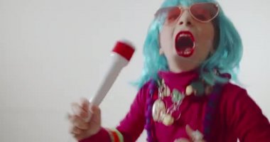 Komik, sevimli, mutlu çocuk mikrofonla şarkı söylüyor. Güneş gözlüğü ve renkli peruk takıp müzik eşliğinde dans ediyor.