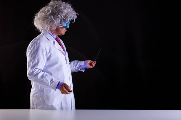 Crazy science teacher in white coat with unkempt hair in funny eye glasses holding a wand to point at the blackboard 