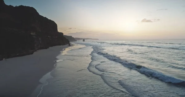 Восход солнца на волнистом берегу океана с высоты птичьего полета. Солнце встает у темной скалы, морская вода умывает песчаный пляж — стоковое фото