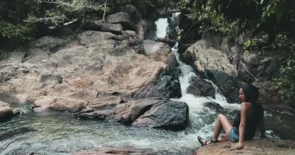 Ταϊλάνδη καταρράκτης ποταμός ζούγκλα: τουριστικός κάθεται σε πετρώδη όχθη ρέμα νερού πτώση και να απολαύσετε της φύσης — Αρχείο Βίντεο