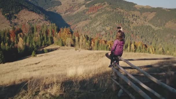 Жінка з мобільним телефоном у гірському ландшафтному повітрі. Красуня сидить на паркані. Палена трава долина — стокове відео