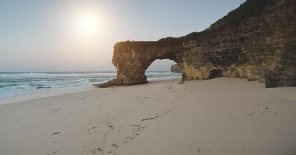 태양은 모래 바다 해안에 있는 절벽 벽에 있는 자연의 구멍을 통해 하늘을 날고 있습니다. 해변의 관광 명소 — 비디오