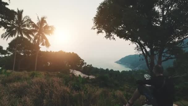 Hombre control drone tiro paisaje de montaña en la luz del atardecer. Adulto joven sentado bajo el árbol — Vídeos de Stock