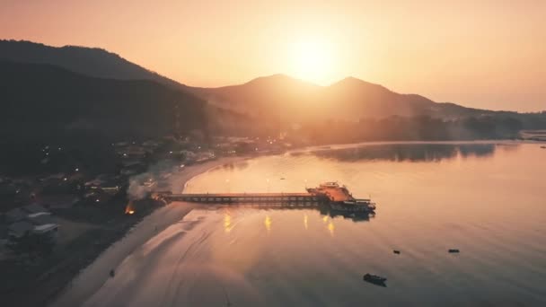 Vuelo lento aéreo sobre la playa de la tarde. Muelle en el mar, casas de luz alrededor de la montaña con árboles, retroiluminación — Vídeo de stock