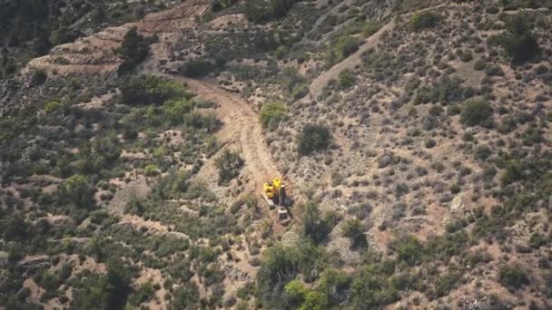 Aerial yellow excavator digs ground in mountains, highway construction. Dangerous work in wilderness — Stock Video