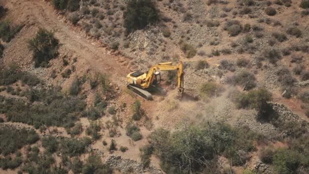 Aeronave uma escavadora cava chão no cume da montanha. Árvores verdes e arbustos em colinas de areia amarela — Vídeo de Stock