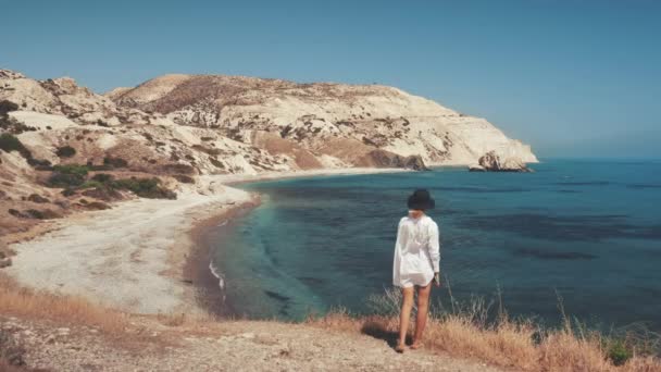 Mujer joven de pie en ropa blanca, mira al océano. Vista trasera trasera. Rocas, isla costera. Viajes — Vídeo de stock