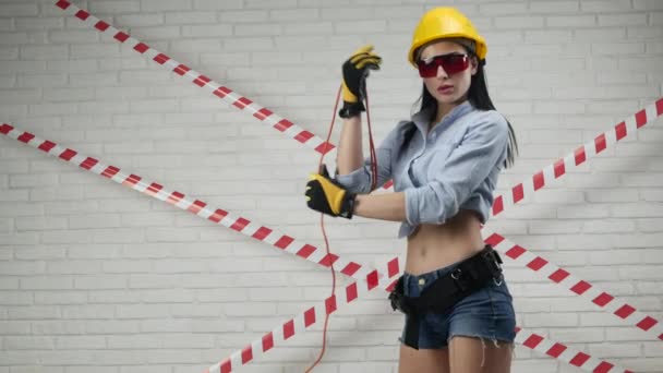 Une femme sexy dans un casque de construction enroule un câble d'alimentation contre un mur de briques blanches — Video