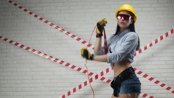 Una donna sexy in un casco da costruzione avvolge un cavo di alimentazione contro un muro di mattoni bianchi — Video Stock