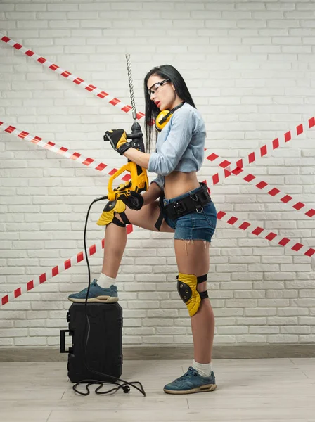 Una donna snella a immagine di un operaio edile con un puncher contro un muro di mattoni con texture bianca — Foto Stock