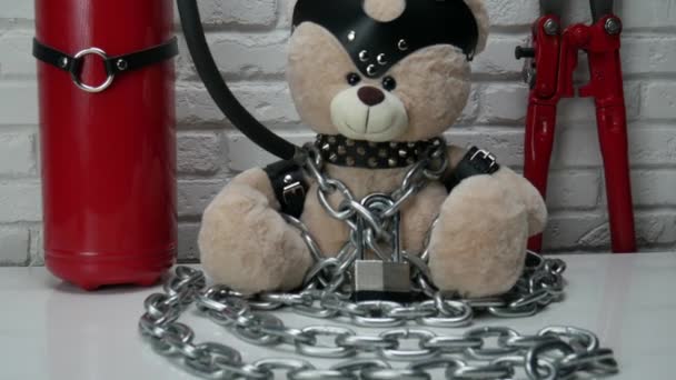 Spielzeug-Teddybär mit Ledergürtel und Maske angekettet und mit Bolzenschneider verriegelt, Zubehör für BDSM-Spiele auf hellem Backsteinhintergrund — Stockvideo