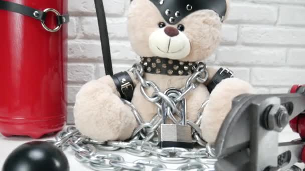 Speelgoed teddybeer gekleed in lederen riemen en een masker geketend en vergrendeld met een betonschaar, accessoires voor BDSM games op een lichte achtergrond textuur van een bakstenen muur — Stockvideo