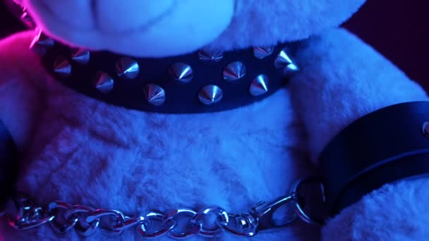 Toy bear dressed in leather belt harness accessory for BDSM games on a dark background in neon light in the smoke — Stock Video