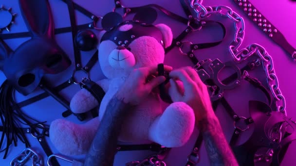 Hände auf einem Spielzeugbär Lederhandschellen auf dem Tisch mit einem BDSM-Accessoire Peitschen Kettenmasken für Spiele in Neonlicht