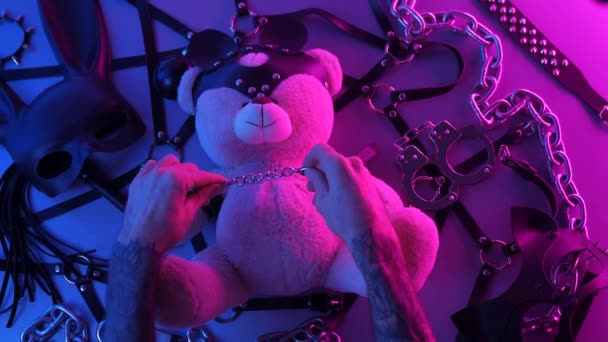 Handen op een speelgoedbeer leren handboeien op tafel met een BDSM accessoire zweep kettingen maskers voor games in neon licht — Stockvideo