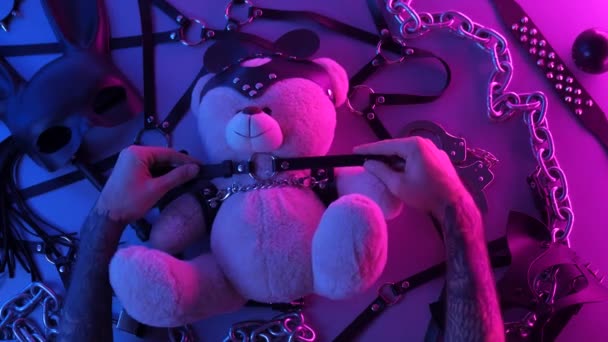 Hände auf einem Spielzeugbär Choker auf einem Tisch mit einem BDSM-Accessoire Peitschen Kettenmasken für Spiele in Neonlicht — Stockvideo