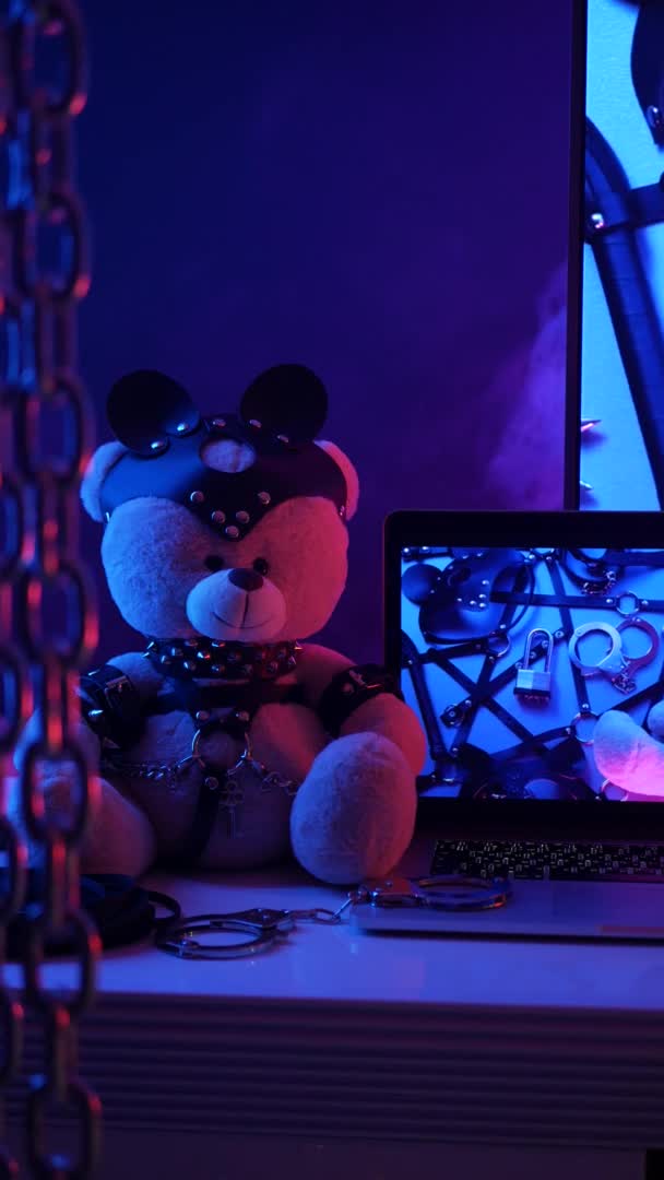 Іграшковий ведмідь у шкіряному аксесуарі для ігор БДСМ поруч з ноутбуком ТБ в неонових кольорах вертикальне відео — стокове відео