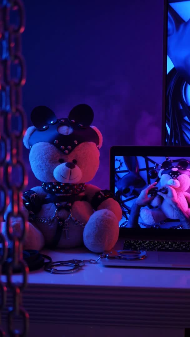 Παιχνίδι αρκούδα σε δερμάτινη ζώνη αξεσουάρ για παιχνίδια BDSM δίπλα σε μια τηλεόραση laptop σε νέον χρώματα κατακόρυφο βίντεο — Αρχείο Βίντεο