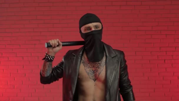 Um criminoso do sexo masculino usando tatuagens uma capa de chuva de couro e uma balaclava com um bastão e uma arma — Vídeo de Stock