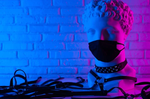 Plaster head of David in BDSM accessories and medical mask from coronavirus whip leather collar and belt in neon light