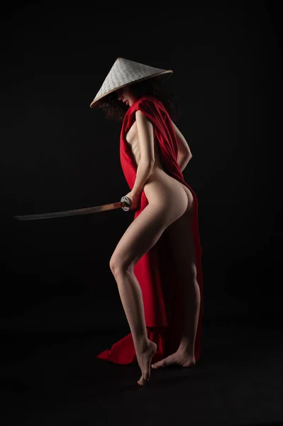 Naken smal kvinna i en röd mantel och en asiatisk hatt med en katana i handen bild av en samuraj — Stockfoto