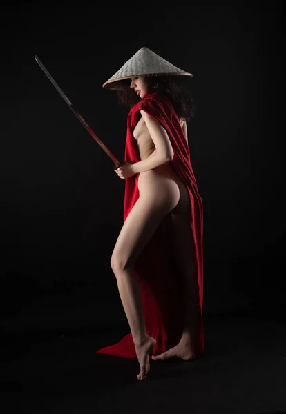 Mulher magra nua em uma capa vermelha e um chapéu asiático com uma katana em sua imagem mão de um samurai — Fotografia de Stock