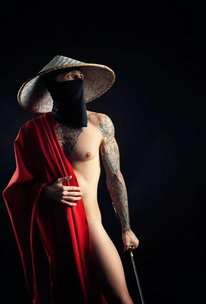 Nahý štíhlý muž samuraj v červené pláštěnce a asijský klobouk s tetovanou katanou — Stock fotografie