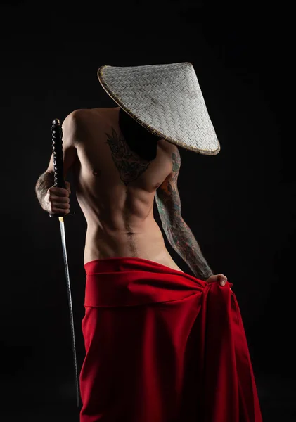 En naken smal man samuraj i röd mantel och en asiatisk hatt med tatuerad katana — Stockfoto