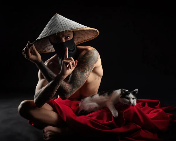 a naked slender samurai man in a red cape and an Asian hat with a cat in his hands
