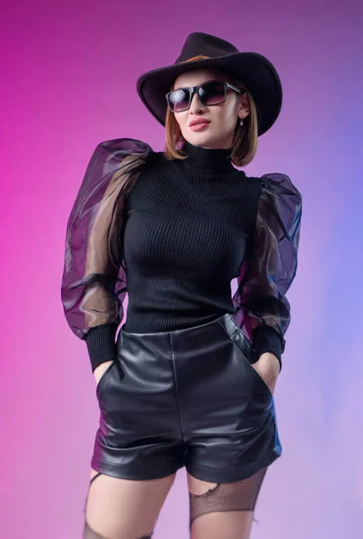 Fashionabla elegant kvinna i svart kläder och hatt poser med neon bakgrund — Stockfoto