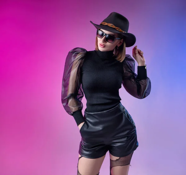 Fashionabla elegant kvinna i svart kläder och hatt poser med neon bakgrund — Stockfoto