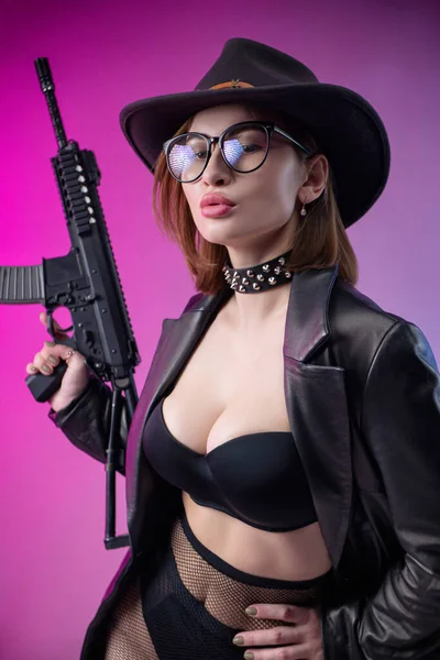 Stylová žena v černém oblečení a klobouk pózuje na neonovém pozadí s airsoftovou pistolí — Stock fotografie
