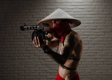 Asya üçgen şapkalı, dövmeli, PPSh-41 havalı, yumuşak tabancalı ve arka planında tuğla duvar olan bir adam.