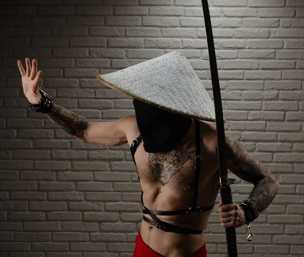 Kırmızı pelerinli bir samuray ve elinde tuğla bir duvarın arkasına dayanmış bir katana kılıcı olan Asyalı üçgen bir şapka. — Stok fotoğraf