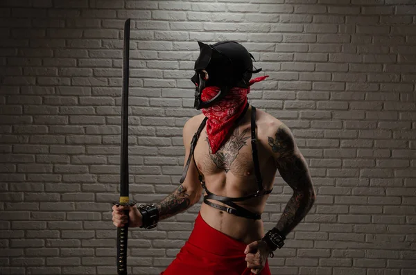 Un hombre samurai con una máscara de demonio con tatuajes y una espada katana en sus manos sobre el fondo de una pared de ladrillo — Foto de Stock